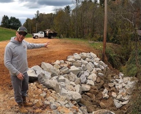 A man stands near a creek where large rocks line the stream to prevent erosion.