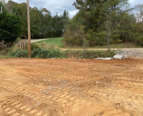 A smooth red dirt lot surrounded by trees and a driveway.